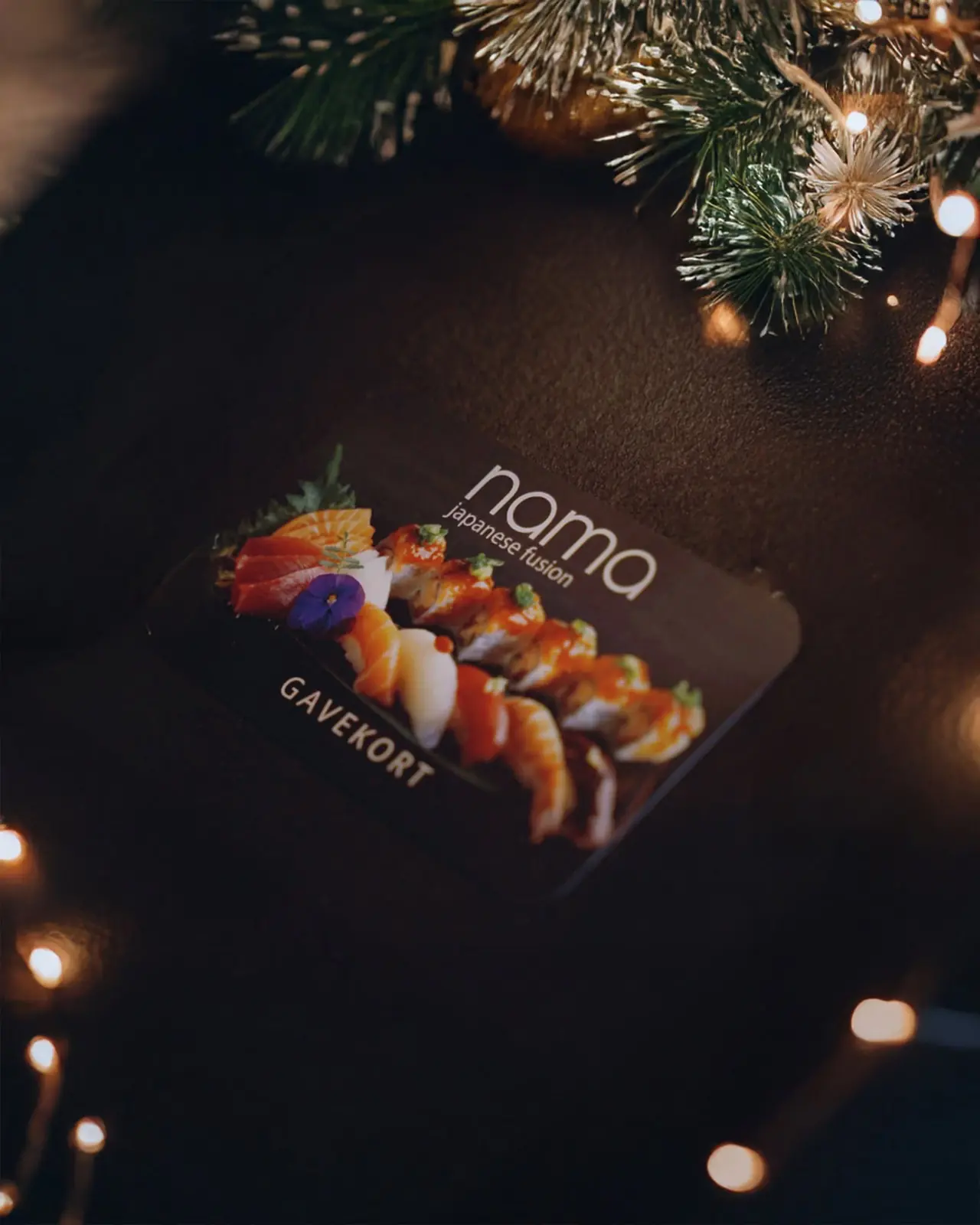 NAMA gift card with sushi artwork on a dark table surrounded by fairy lights and pine