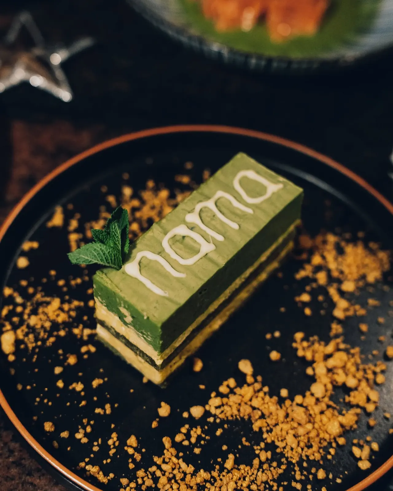 Matcha cake with the NAMA logo piped in white, served with crumble on a dark plate