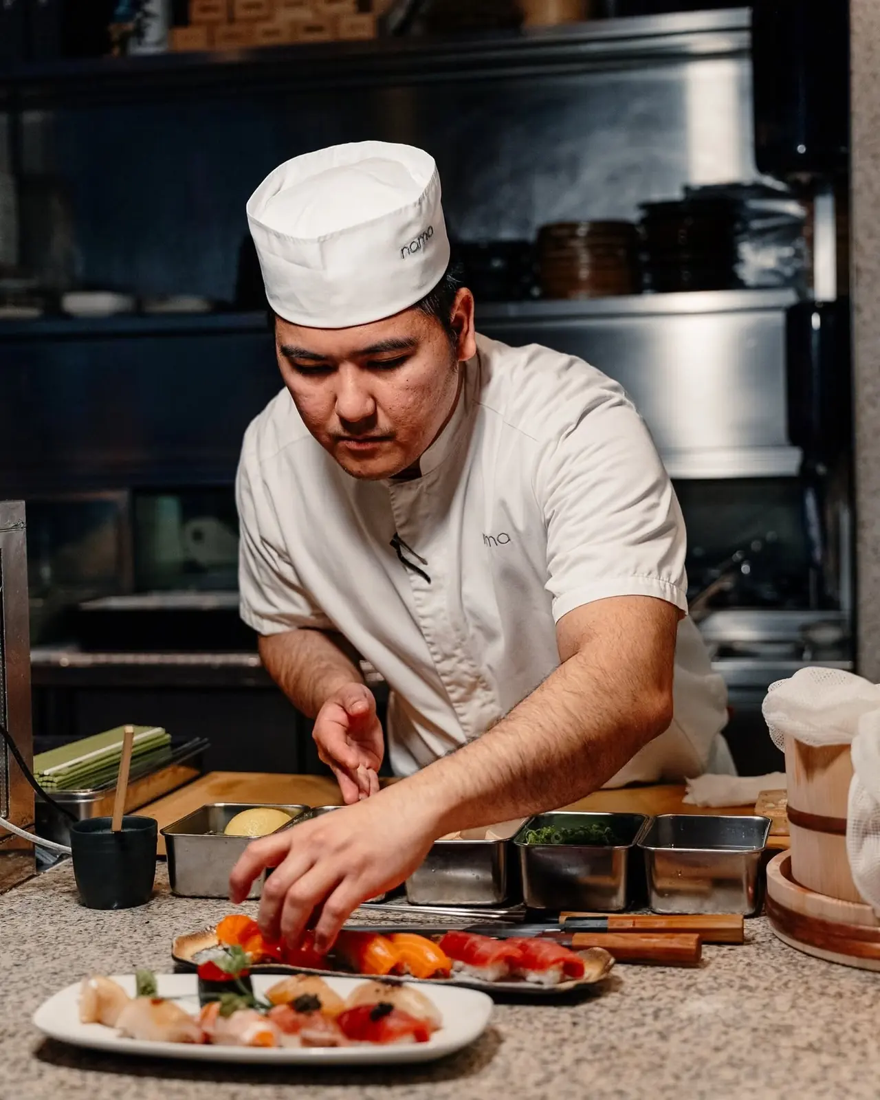Sushi chef in NAMA whites plating nigiri behind the counter