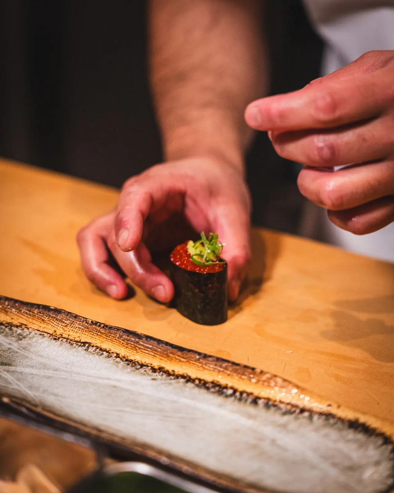 Sushi chef finishing an ikura gunkan by hand on a wooden counter